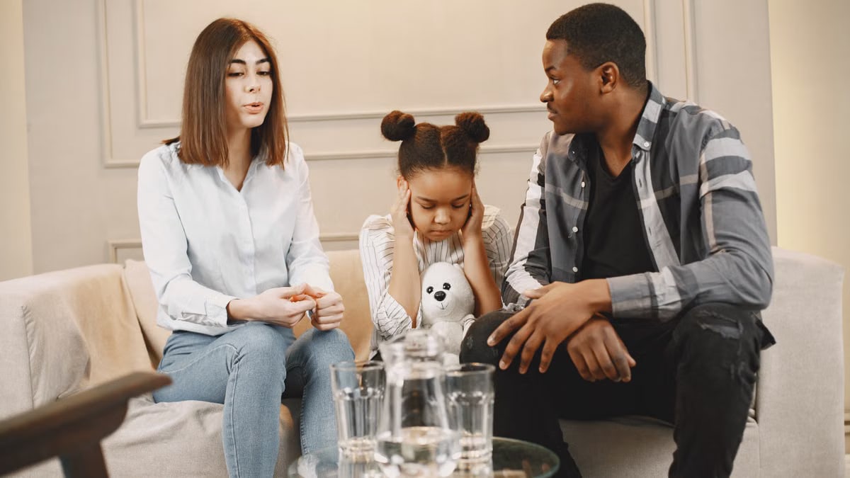 Parents talking to daughter covering ears on sofa.